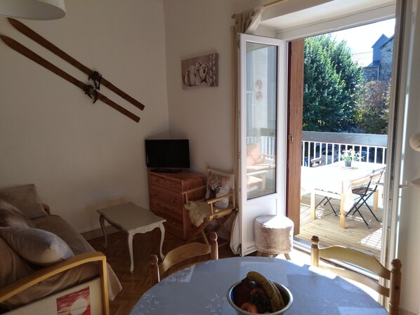 A cozy room with a wooden TV stand, a dining table with a fruit bowl, a white chair, and a view of a balcony with outdoor furniture.