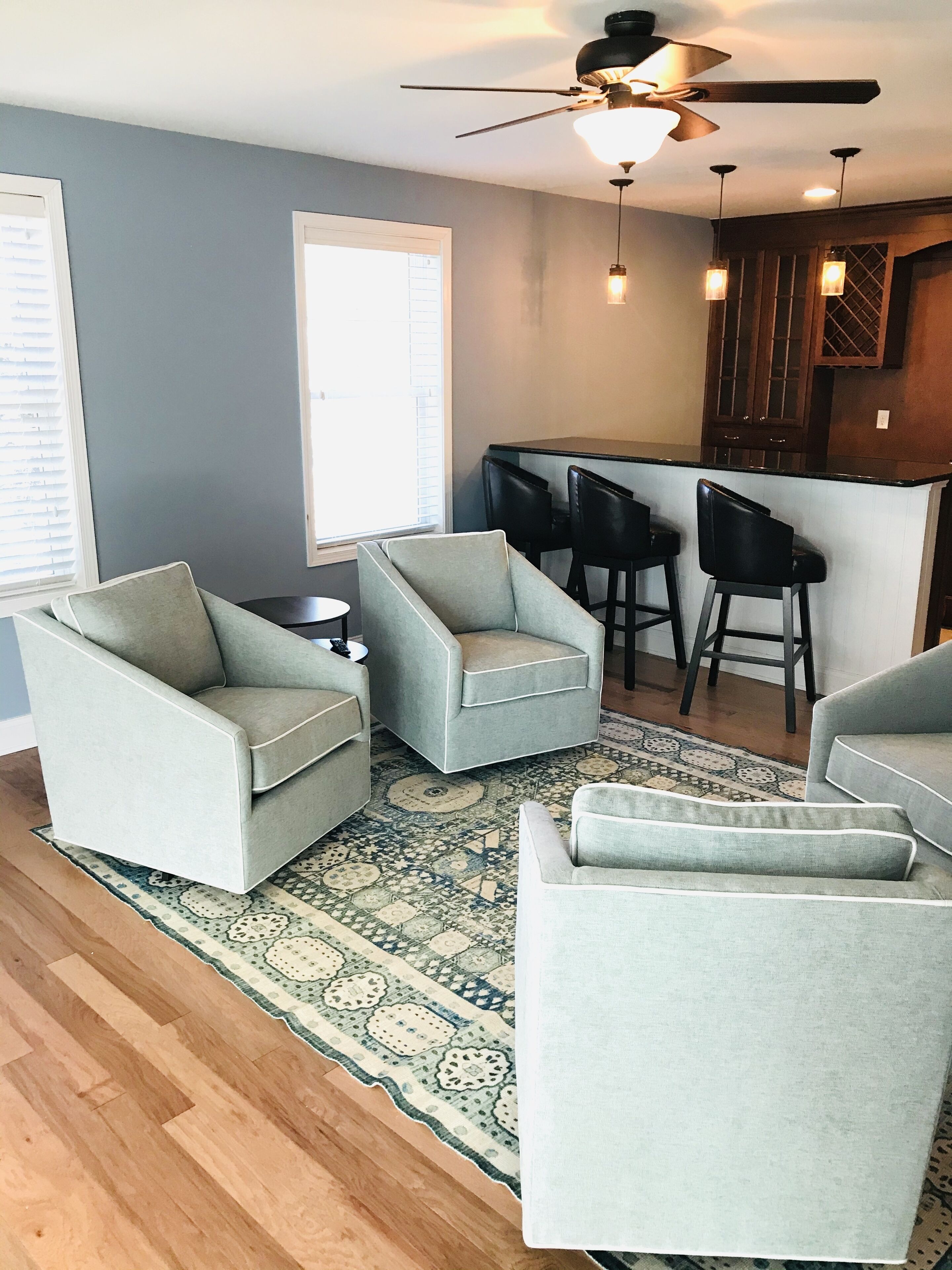 A modern living room with grey armchairs, a coffee table, and a patterned rug. There is a kitchen area with dark wooden cabinets and a bar counter.