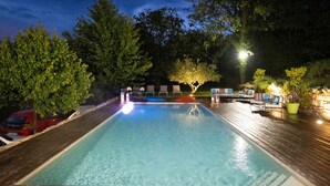 A rectangular swimming pool with a wooden deck surrounding it, a tree in the center, and a red car parked nearby.