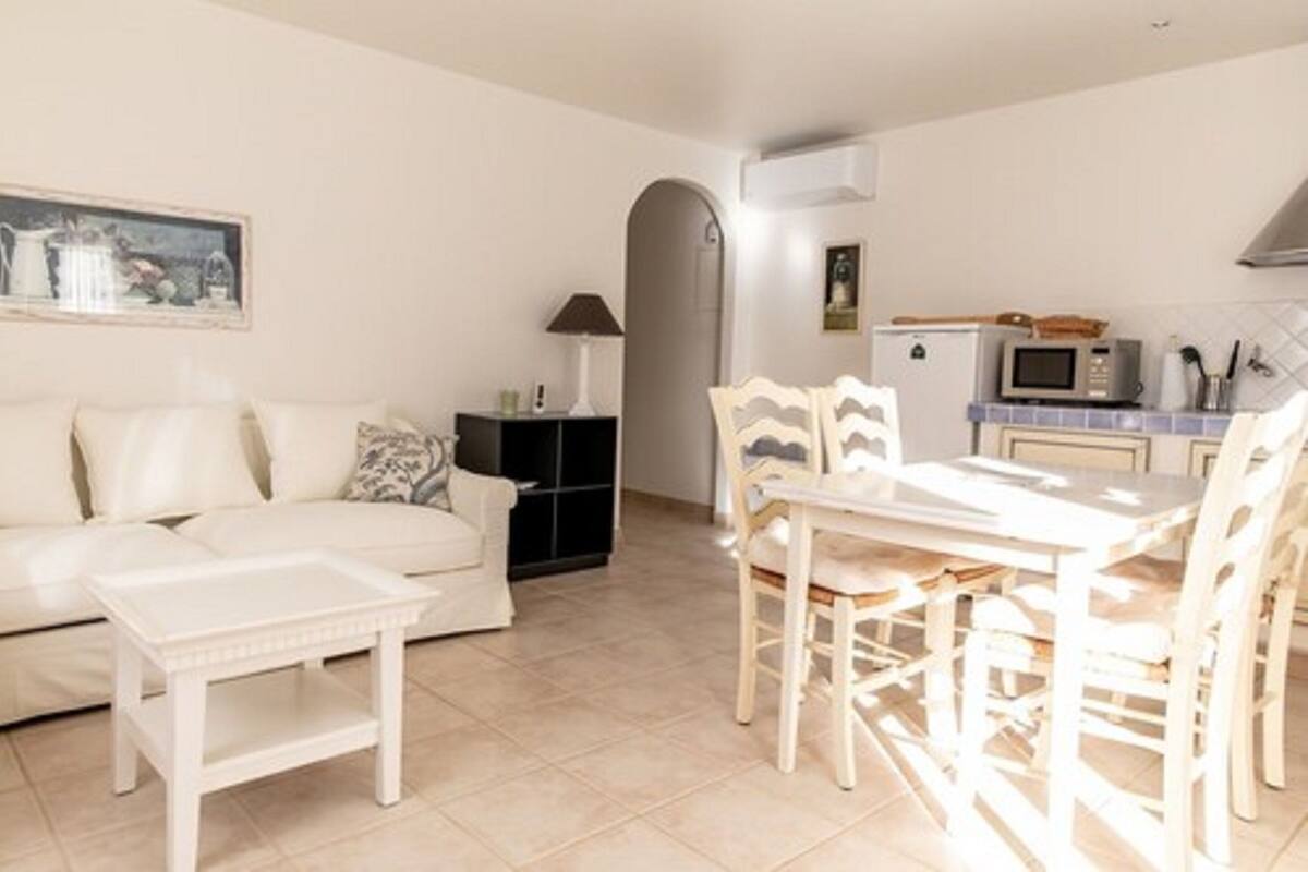 Een ruimtelijke woonkamer met een witte bank, een eettafel met stoelen, een kitchenette met een magnetron en een zwart kastje.
