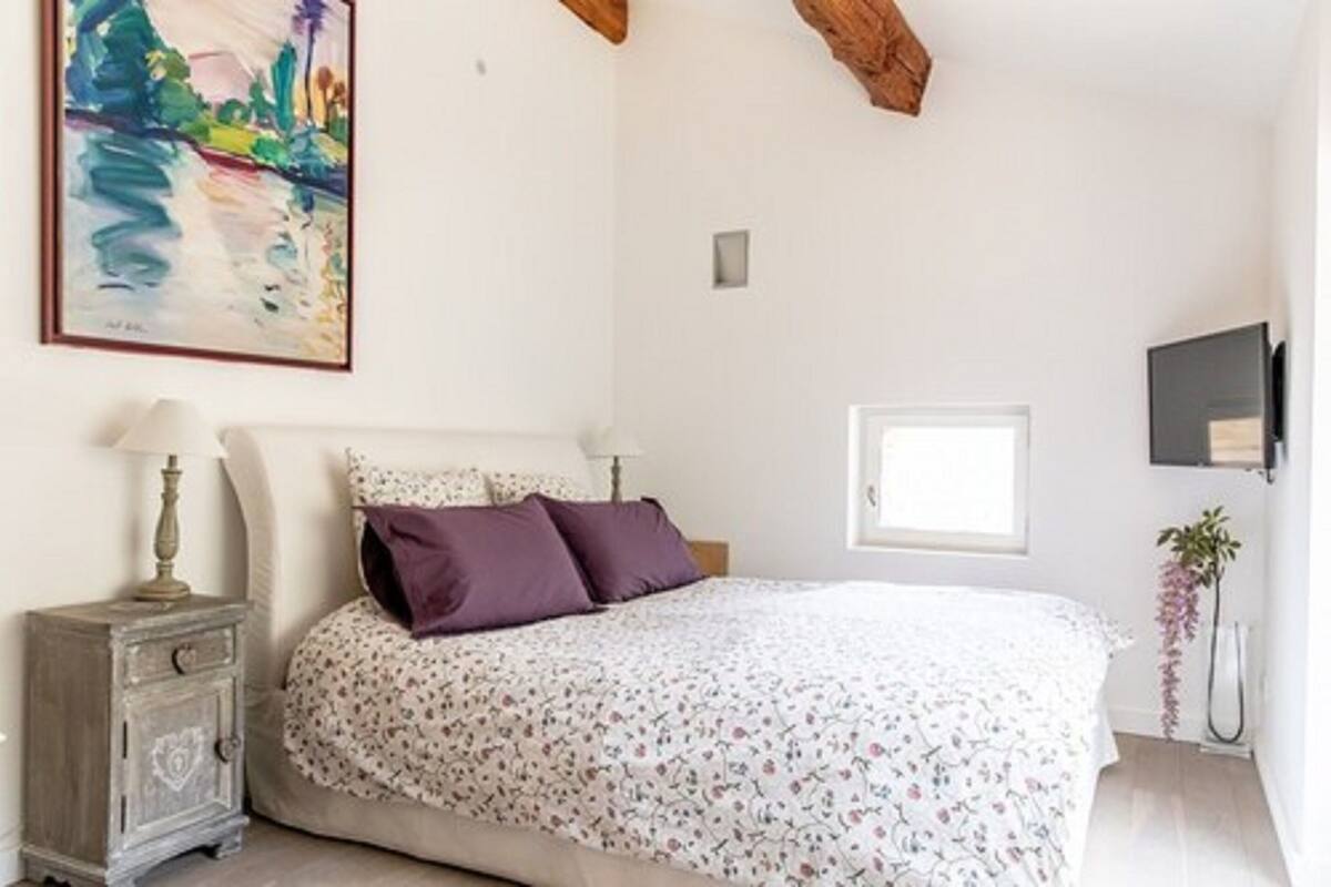 Een slaapkamer met een bed, nachtkastje, schilderij, raam en een plant.