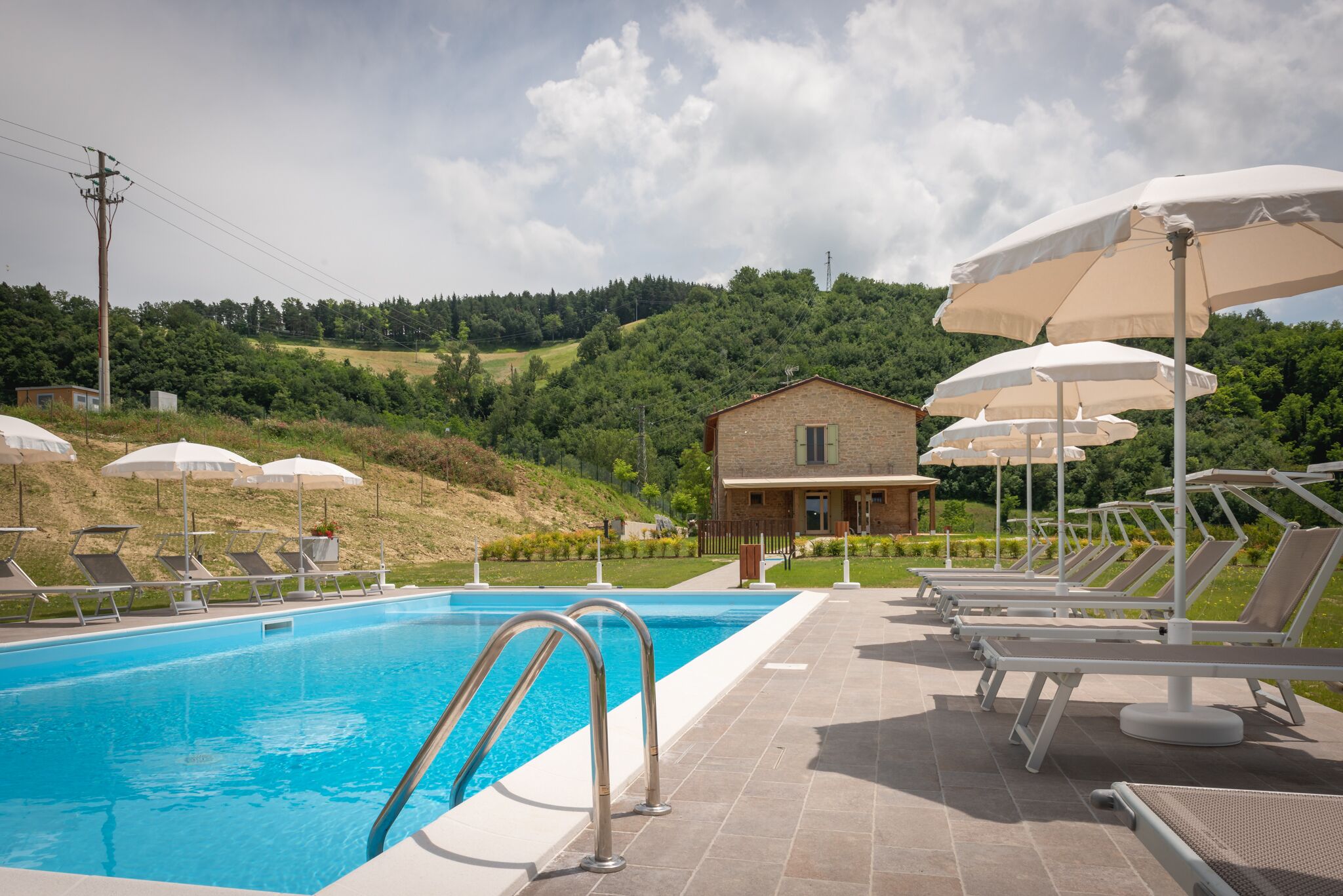 A swimming pool with lounge chairs and umbrellas, a small building, and a hilly landscape in the background.