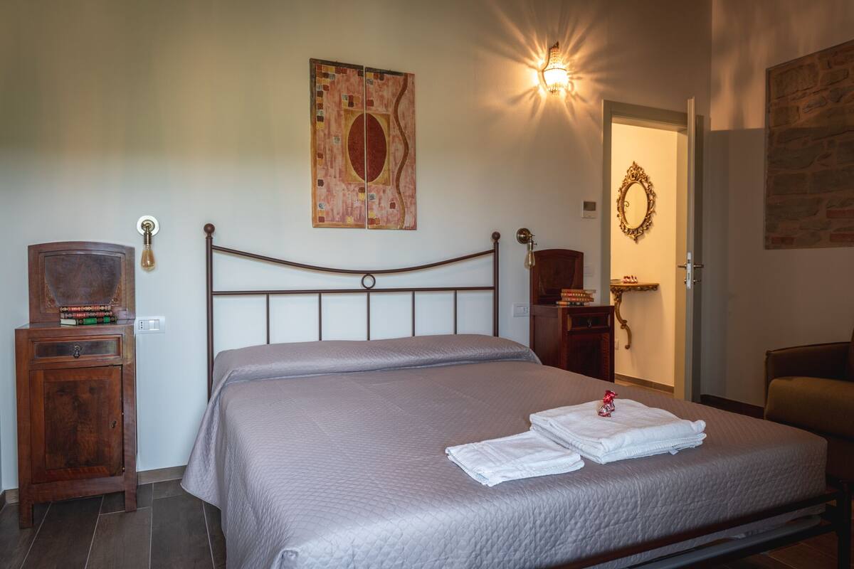 Un lit bien fait, avec une tête de lit, une table de chevet avec une lampe, une œuvre d’art accrochée au mur et une porte ornée d’une guirlande décorative.