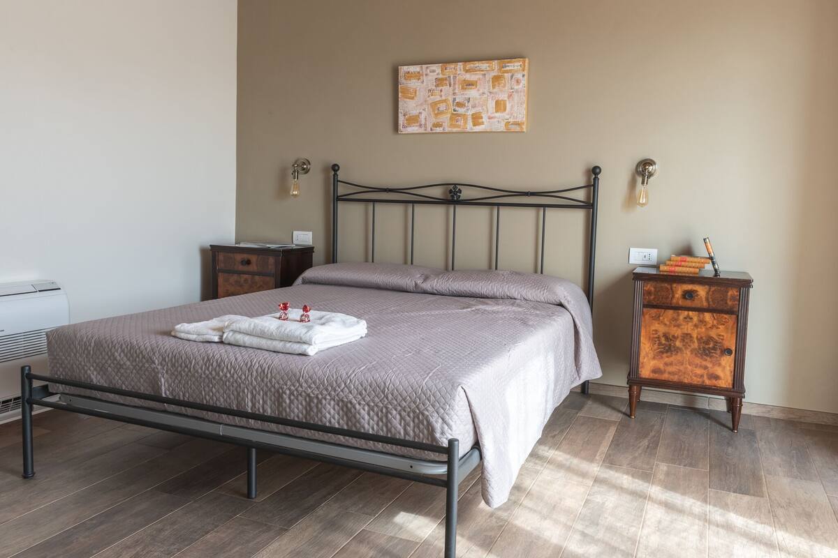 Un lit bien fait, recouvert d’une courtepointe, une table de chevet avec une lampe et une petite armoire contenant des objets décoratifs.