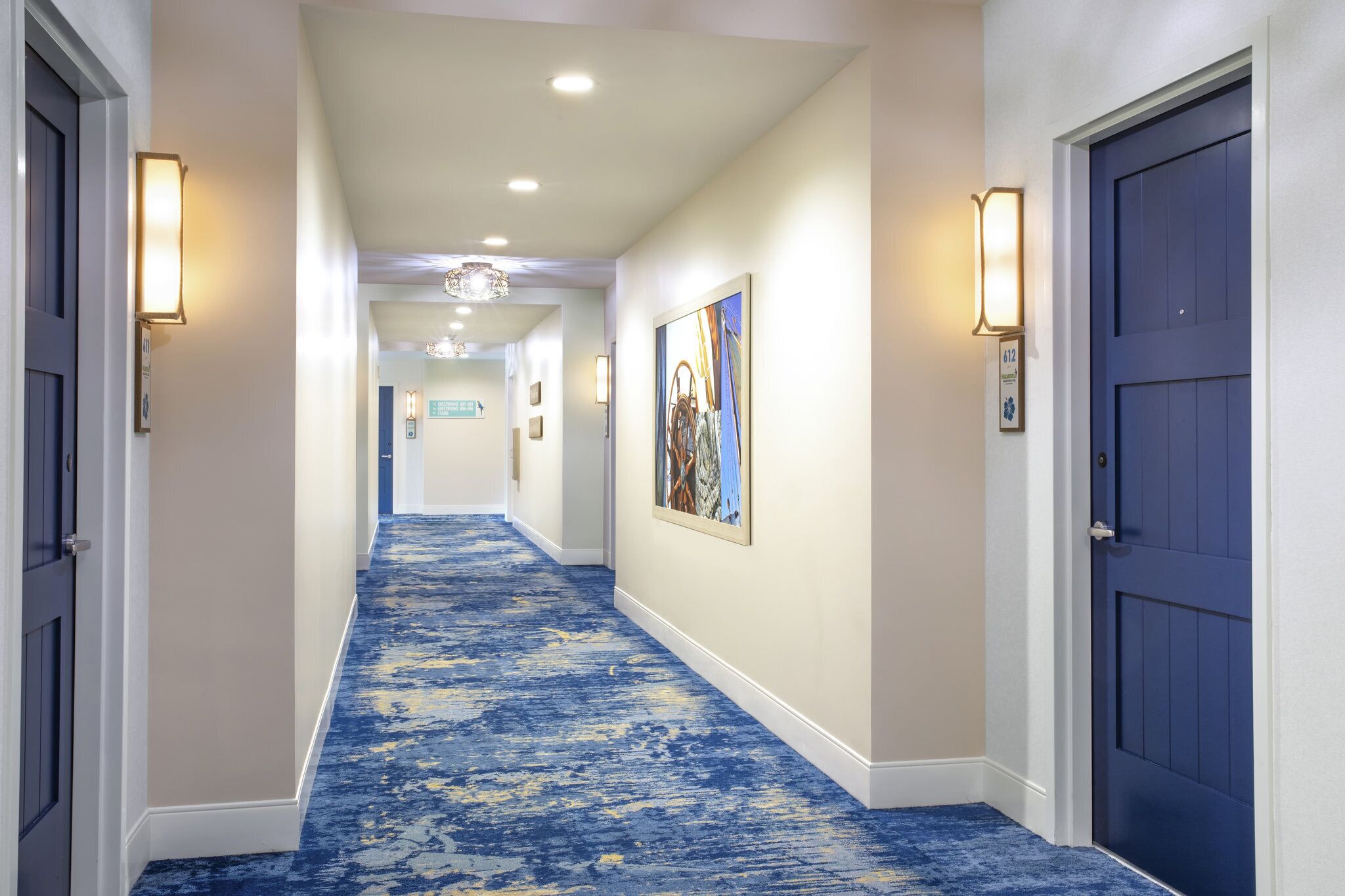Un pasillo de hotel con alfombra azul, paredes blancas y puertas azul oscuro.