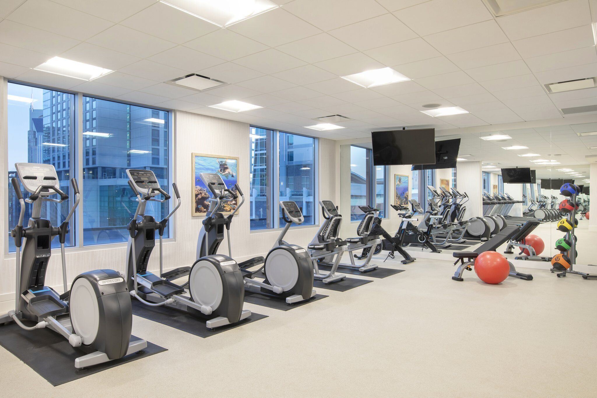 A gym with various cardio machines, exercise balls, and a large window with a city view.