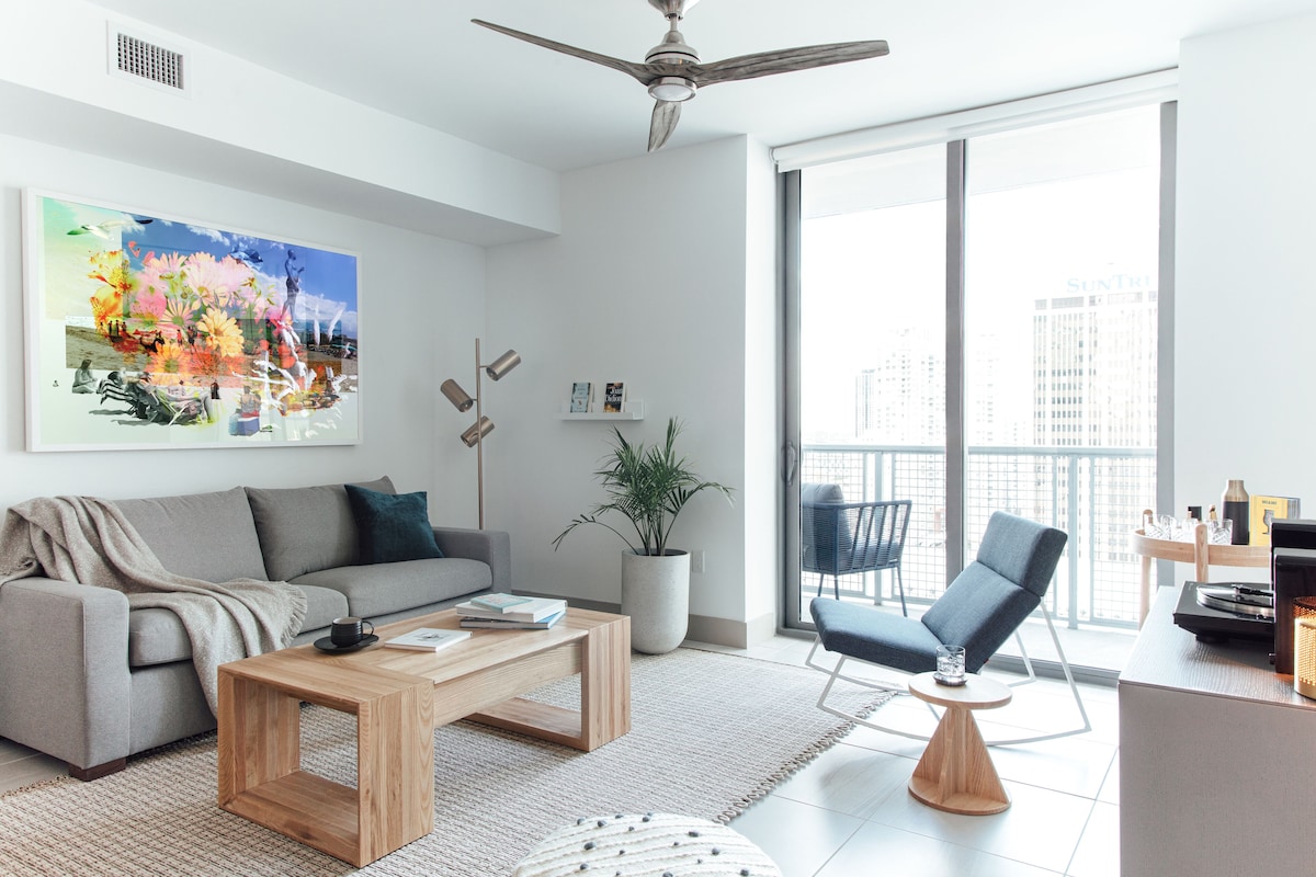 A modern living room with a sofa, coffee table, and armchair, a large window with a city view, and a ceiling fan.