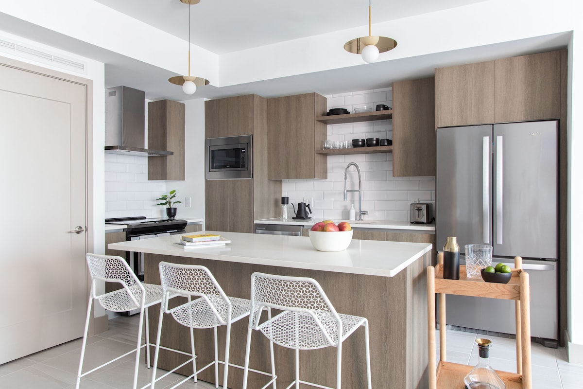 A modern kitchen with a central island, bar stools, stainless steel appliances, and wooden cabinetry.
