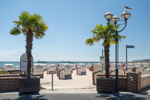 Acceso a la playa con palmeras, un cartel de sanitarios y vista al mar y un muelle al fondo.