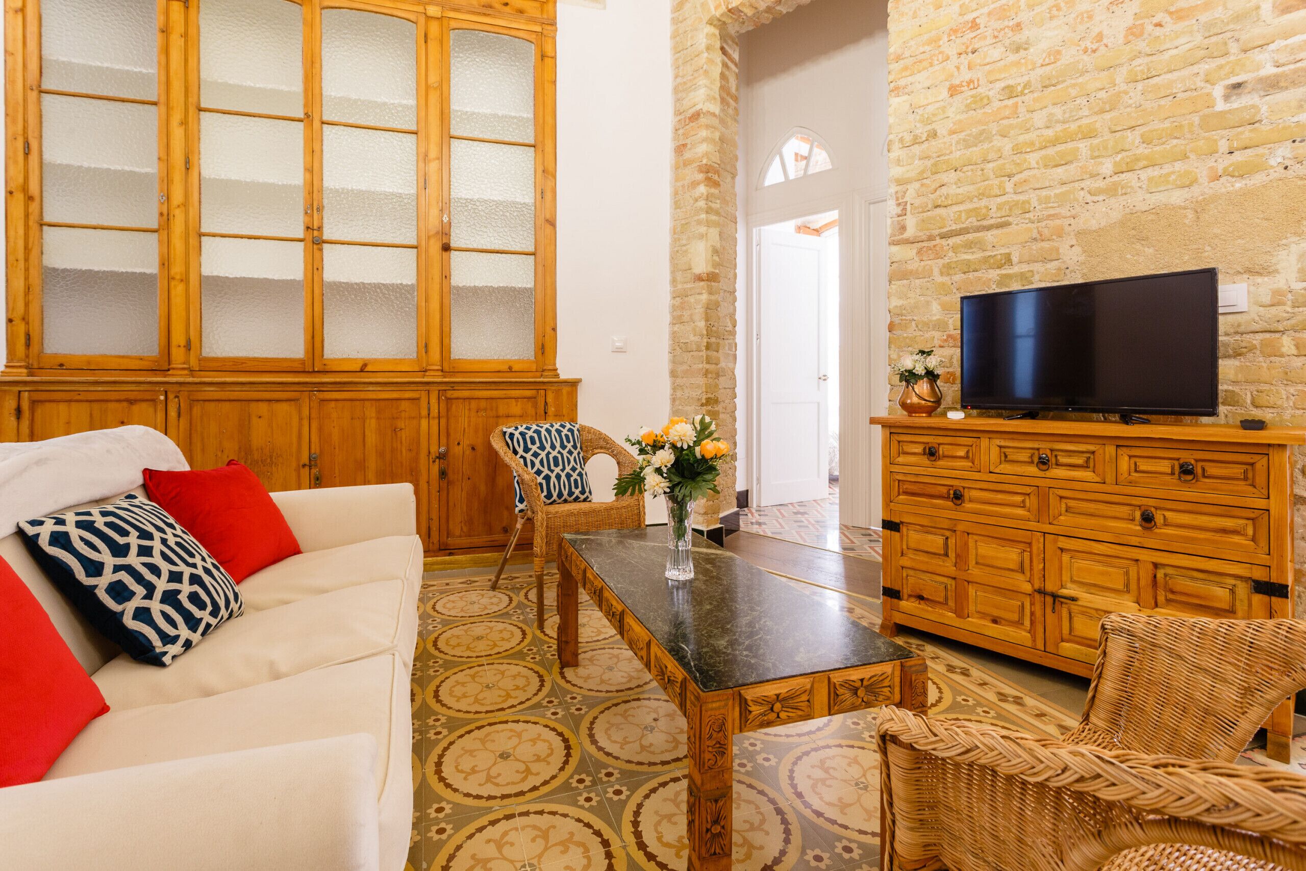 A living room with a white sofa, red and blue patterned pillows, a wooden cabinet with glass doors, a television on a wooden stand, and a brick wall.