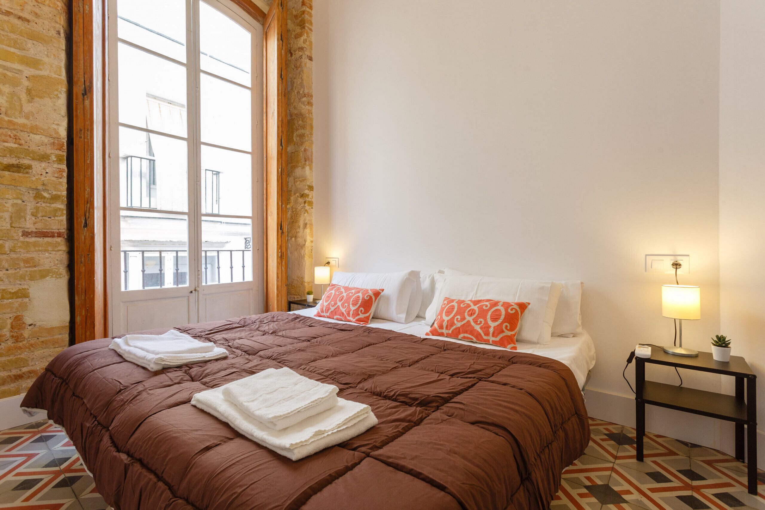 A hotel room with a large bed, a wooden-framed window, a small table with a lamp, and a potted plant.
