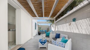 Een overdekt terras met witte rieten meubels, een blauwe mat en een bloempot.