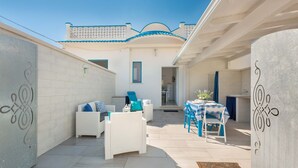 A patio area with white wicker furniture, a table with a blue and white checkered tablecloth, and a kitchen visible through an open door. - 1688 Villa Blu Cobalto - Appartamento 2 (San Pietro in Bevagna)