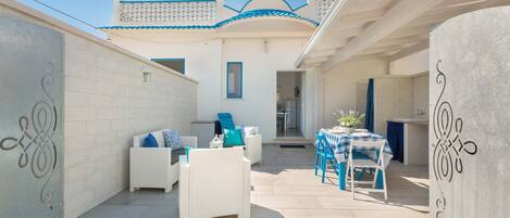 A patio area with white wicker furniture, a table with a blue and white checkered tablecloth, and a kitchen visible through an open door.