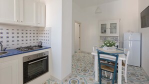 A kitchen with white cabinets, a gas stove, an oven, and a dining area with a table and chairs. - 1688 Villa Blu Cobalto - Appartamento 2 (San Pietro in Bevagna)