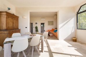 A modern living room with a glass dining table, white chairs, a wooden cabinet, and a sofa with colorful cushions.