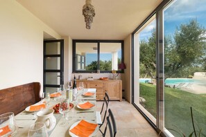 A dining area with a table set for four, a wooden bench, and a view of a swimming pool and greenery outside.