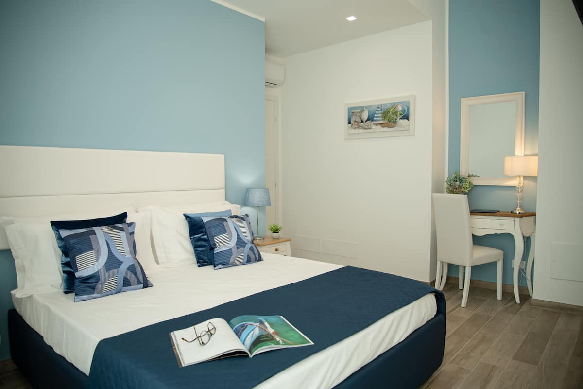 Ein ordentlich bezogenes Bett mit blauer Bettdecke und weißen Kissen, ein Nachttisch mit Lampe sowie ein Schreibtisch mit Stuhl in einem Zimmer mit hellblauen Wänden.