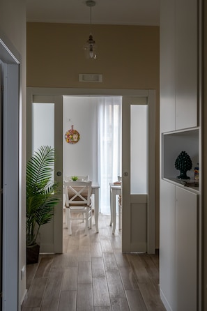 A hallway with a wooden floor, white walls, a ceiling light, a potted plant, and a dining area with a table and chairs.