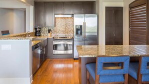 Una cocina compacta con un refrigerador de acero inoxidable, un horno empotrado y una cafetera. Hay una mesa de comedor de madera con sillas tapizadas en azul.