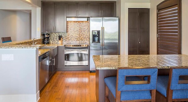 A compact kitchen with a stainless steel refrigerator, a built-in oven, and a coffee maker. There is a wooden dining table with blue upholstered chairs. - Close to SeaWorld Spacious Two Bedroom w/ pools, hot tubs, bbq pits and family adventures (Orlando)