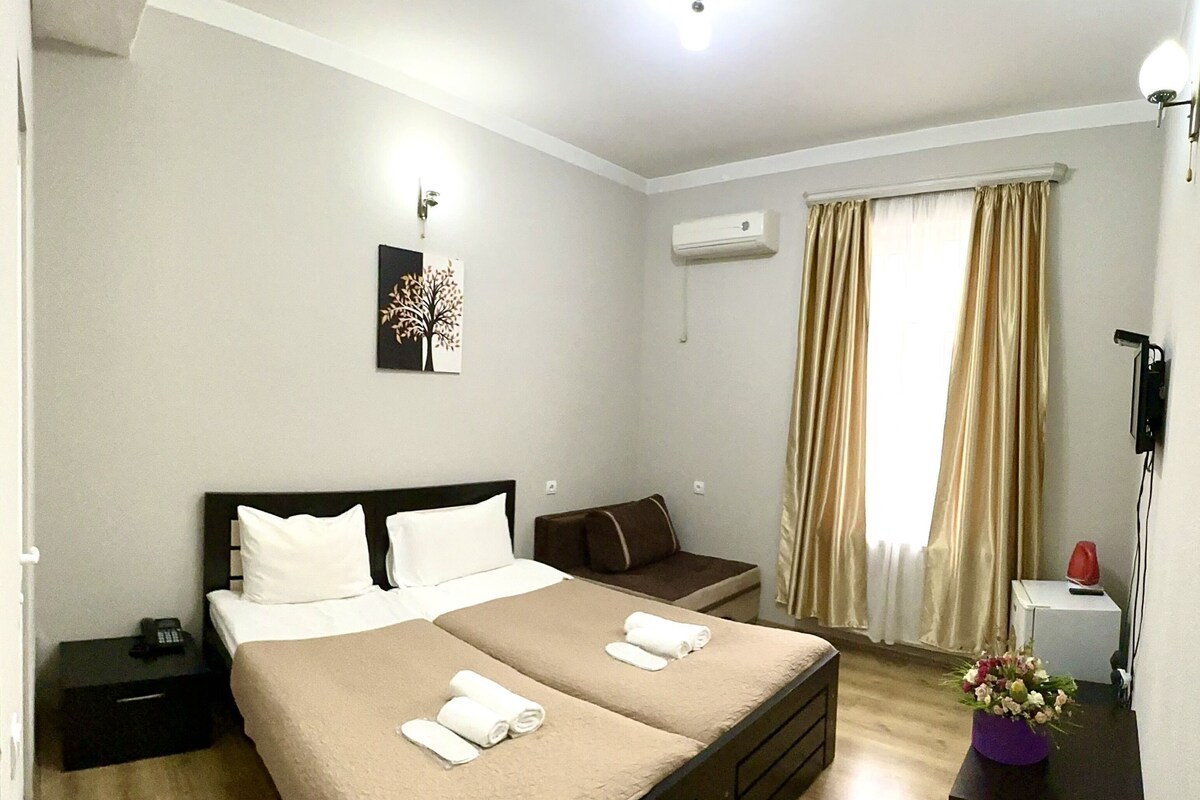 Een hotelkamer met een tweepersoonsbed, nachtkastjes, een kleine bank, een airconditioning en een raam met gordijnen, plus een bedlampje.