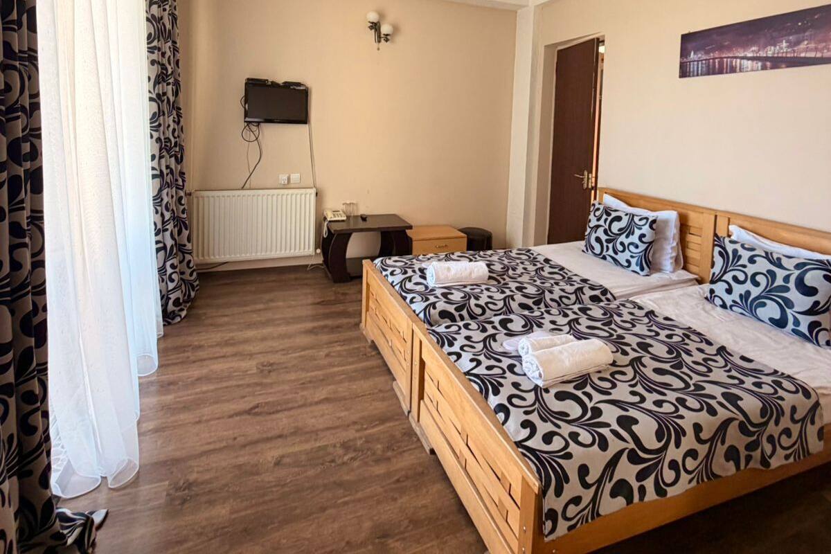 Un dormitorio con una cama de madera, ropa de cama estampada, un escritorio con silla, un radiador, un televisor montado en la pared y una puerta.
