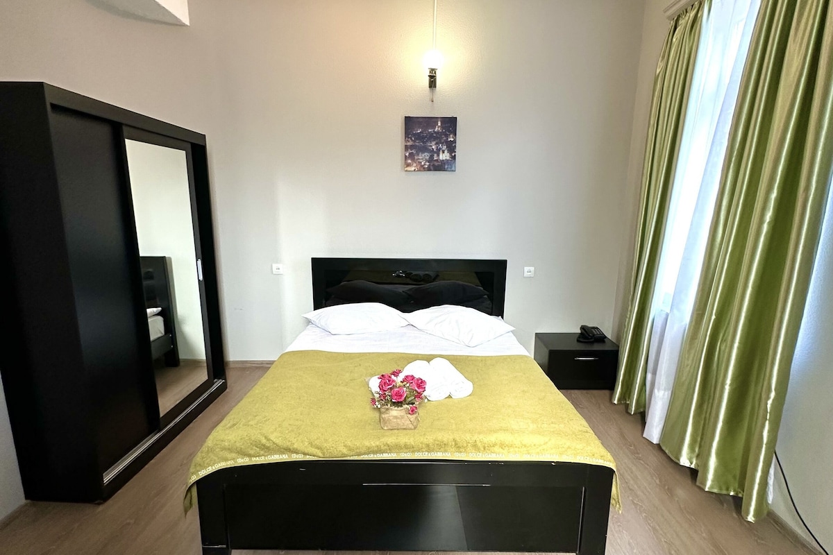 Een slaapkamer met een bed, nachtkastje, spiegel en raam met gordijnen.