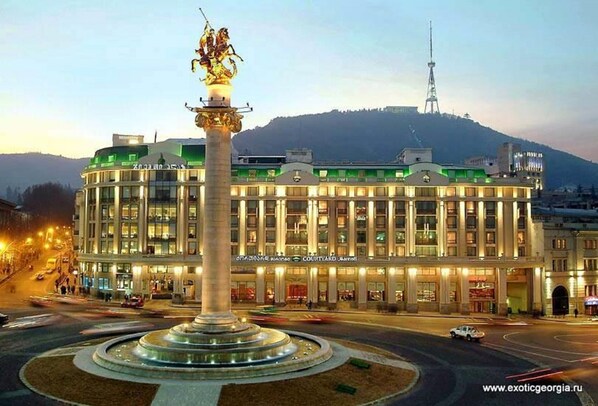 A hotel with a central fountain, a large building with multiple floors, and a tower in the background. - Qeroli old town (Tbilisi)