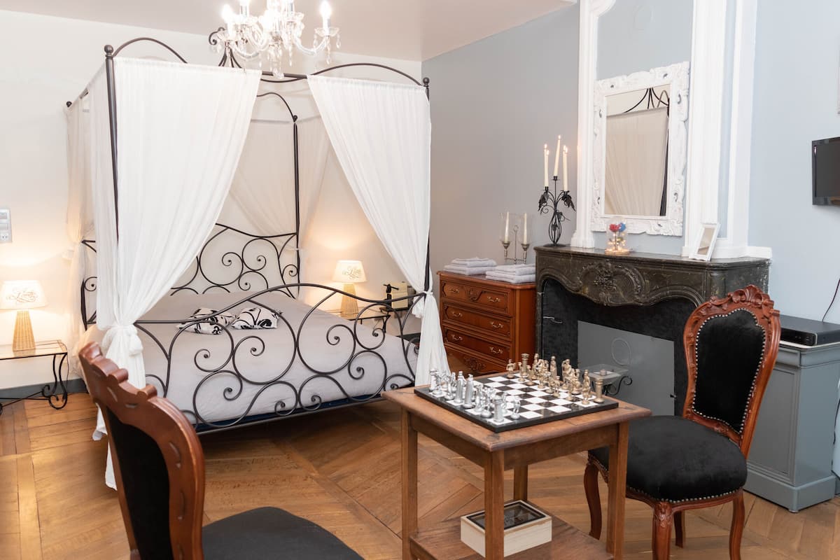 A bedroom with a four-poster bed, a wooden chair, a chess table, and a mirror above a fireplace.