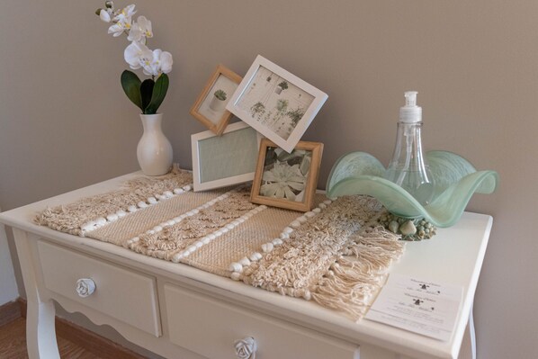 Comoda branca com um corredor de macramé, impressões botânicas emolduradas, um vaso branco com flores brancas e uma garrafa transparente com bomba.