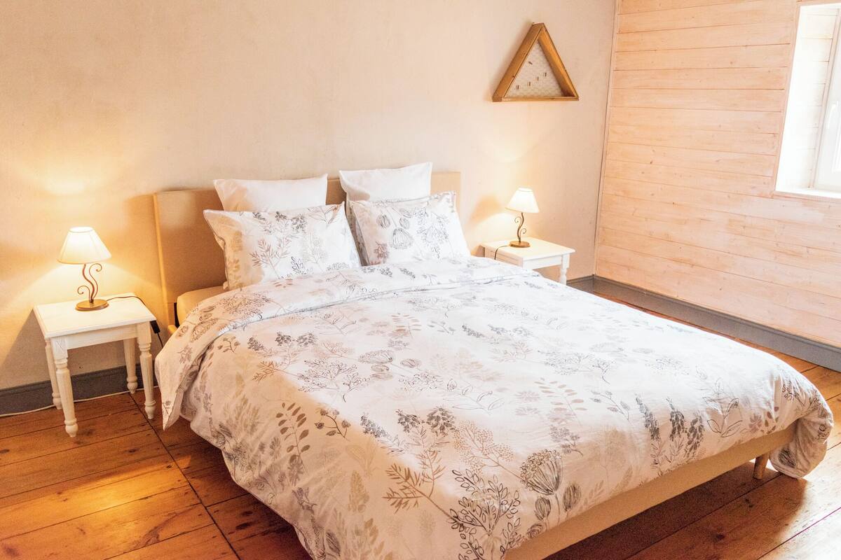 A bedroom with a wooden floor, a bed with floral bedding, two bedside tables with lamps, and a wooden wall panel.