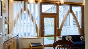 A modern kitchen with wooden cabinets, a sink, and a dining area with a table and chairs. There are large windows with vertical blinds, a wooden bench, and a staircase leading to an upper level. - The Two Arches - Les Arches de Muschamp - Holiday Homes with Style & Comfort (Cussay, Centre-Val De Loire)