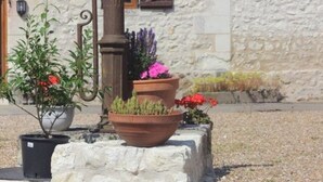 A stone building with a tiled roof, a wooden door, and a window with a wooden shutter. There are potted plants with flowers in front of the building. - The Two Arches - Les Arches de Muschamp - Holiday Homes with Style & Comfort (Cussay, Centre-Val De Loire)