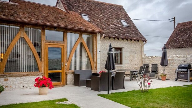 A stone building with a wooden door and windows, outdoor seating with umbrellas, and a barbecue grill. - The Two Arches - Les Arches de Muschamp - Holiday Homes with Style & Comfort (Cussay, Centre-Val De Loire)