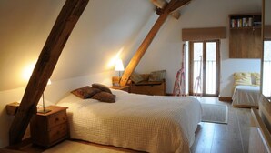 A bedroom with a sloped ceiling, wooden beams, a bed with a white bedspread, a nightstand, a lamp, a chair, and a bookshelf. - The Two Arches - Les Arches de Muschamp - Holiday Homes with Style & Comfort (Cussay, Centre-Val De Loire)