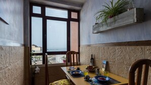 Un coin repas avec une table dressée pour deux personnes, une vue sur l’océan par de grandes fenêtres et une étagère en bois ornée d’une plante.