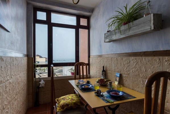 A dining area with a table set for two, a view of the ocean through large windows, and a wooden shelf with a plant.