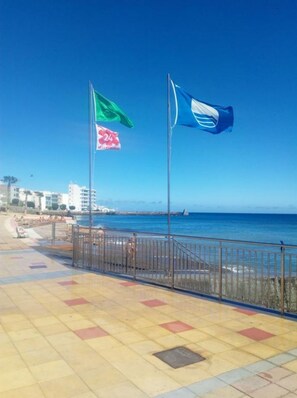 Une promenade en bord de mer avec une allée dallée, une rambarde en métal et deux drapeaux sur des mâts.