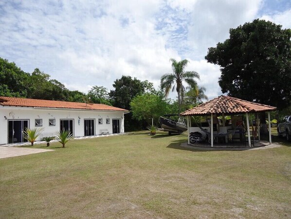 A white building with a red roof, a covered outdoor seating area, and a palm tree in the background. - Pousada Camping e Pesca Bom Abrigo (Ilha Comprida)