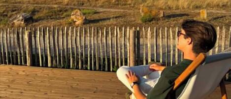 Una persona sentada en una terraza de madera con vista a una playa, con una valla de madera al frente.