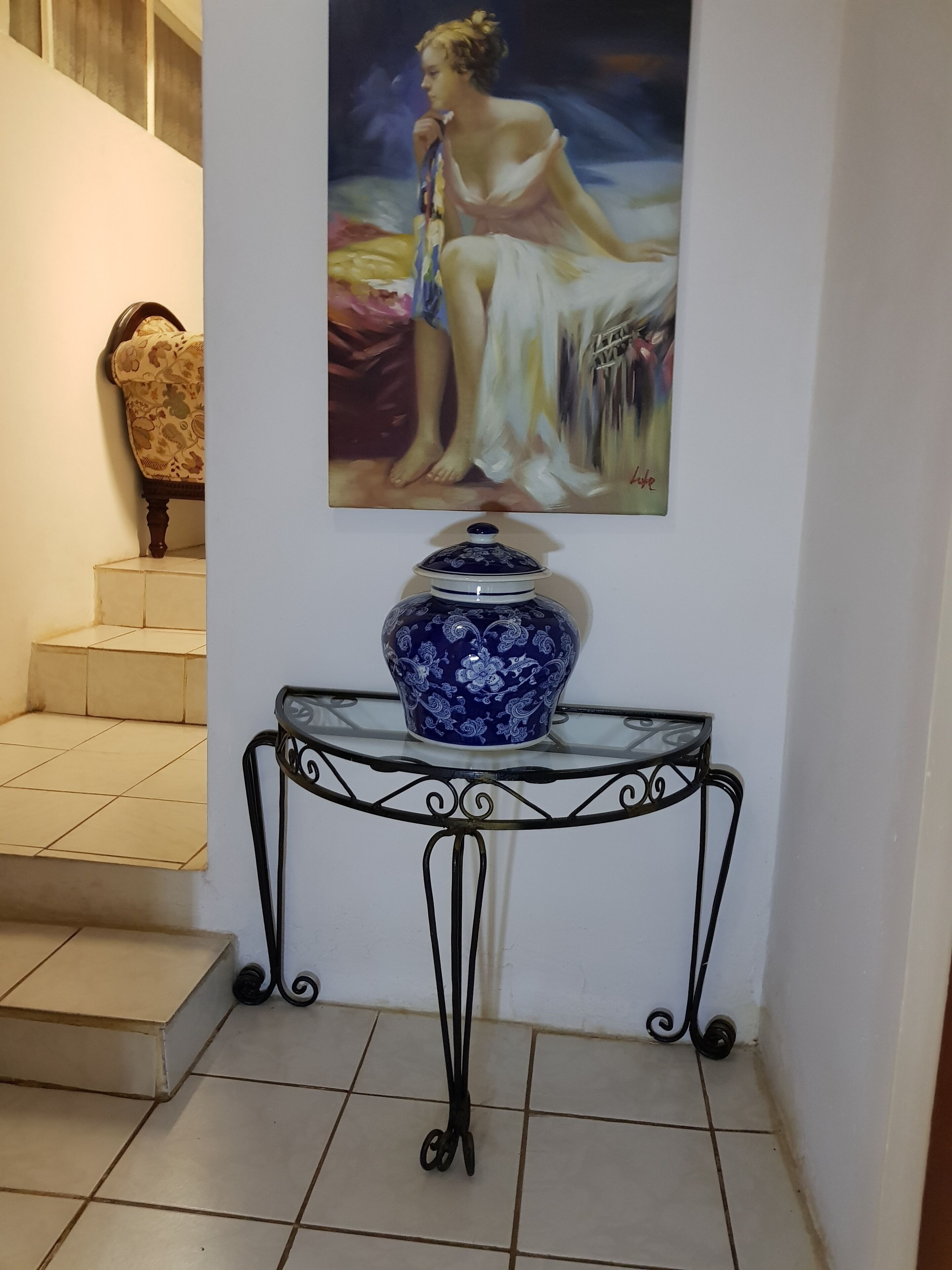 A blue and white ceramic vase on a glass table with metal legs, a painting of a woman on the wall, and a patterned chair in the background.