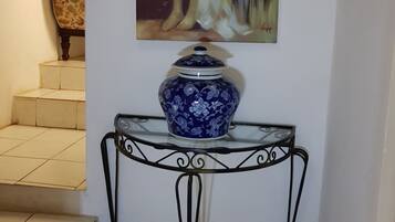 A blue and white ceramic vase on a glass table with metal legs, a painting of a woman on the wall, and a patterned chair in the background.