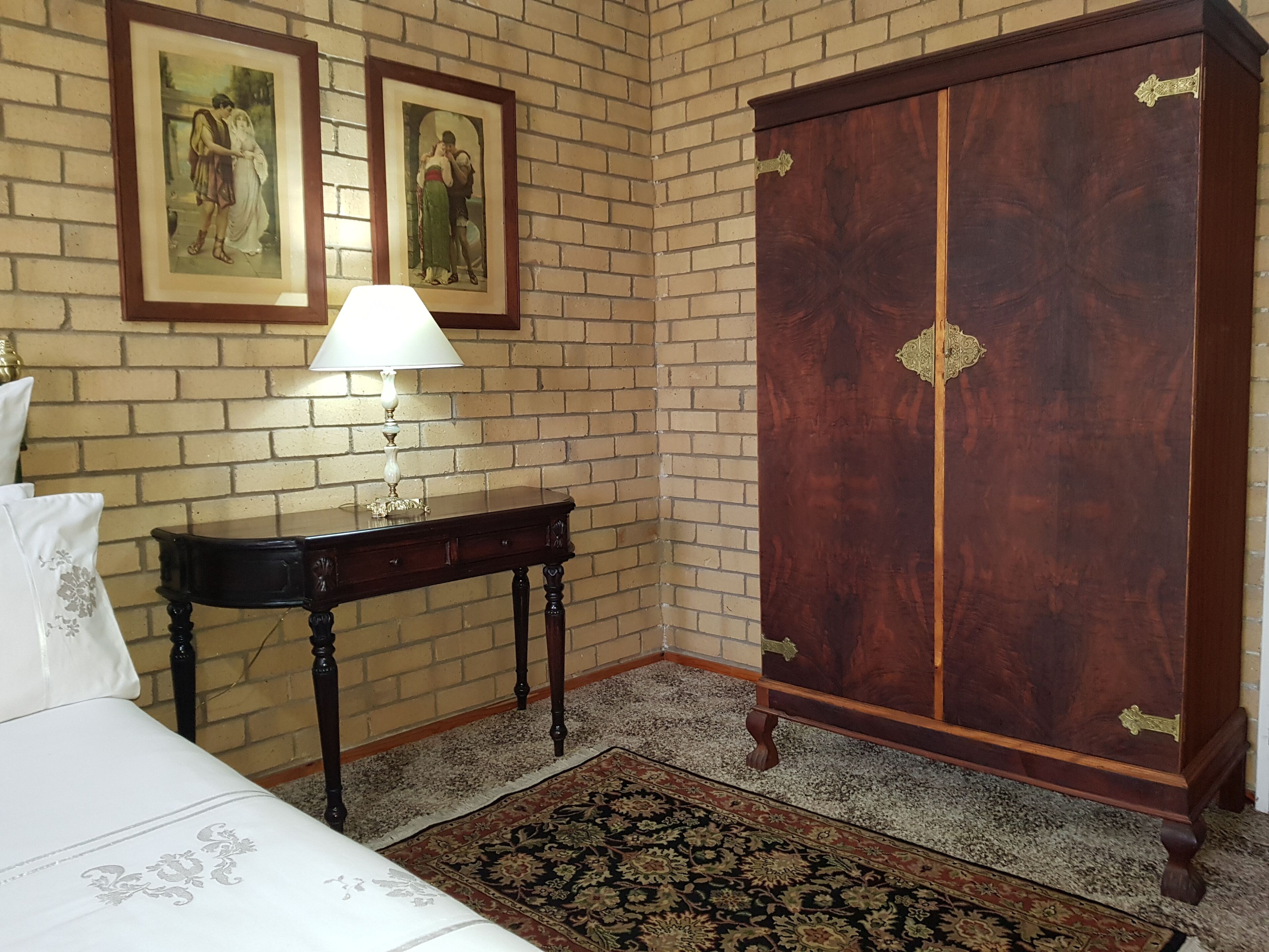 Een kamer met een bed, een tafel met een lamp, een grote houten garderobe en twee ingekaderde foto's aan de muur.