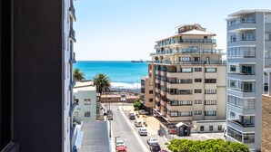 A coastal view from a balcony overlooking a street with cars and buildings, including a multi-story hotel with balconies and a beach in the background. - Bradway Apartments (Cape Town)