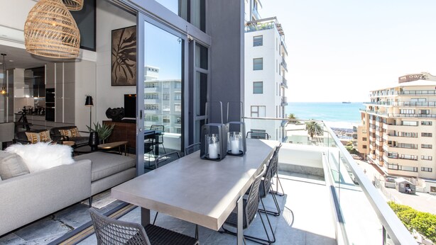 A modern living room with a sofa, coffee table, and dining table with chairs. There is a large window with a view of the ocean and buildings. - Bradway Apartments (Cape Town)