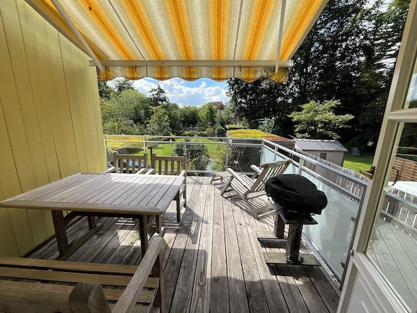 A wooden deck with a table and chairs, a grill, and a view of a garden and house in the background.