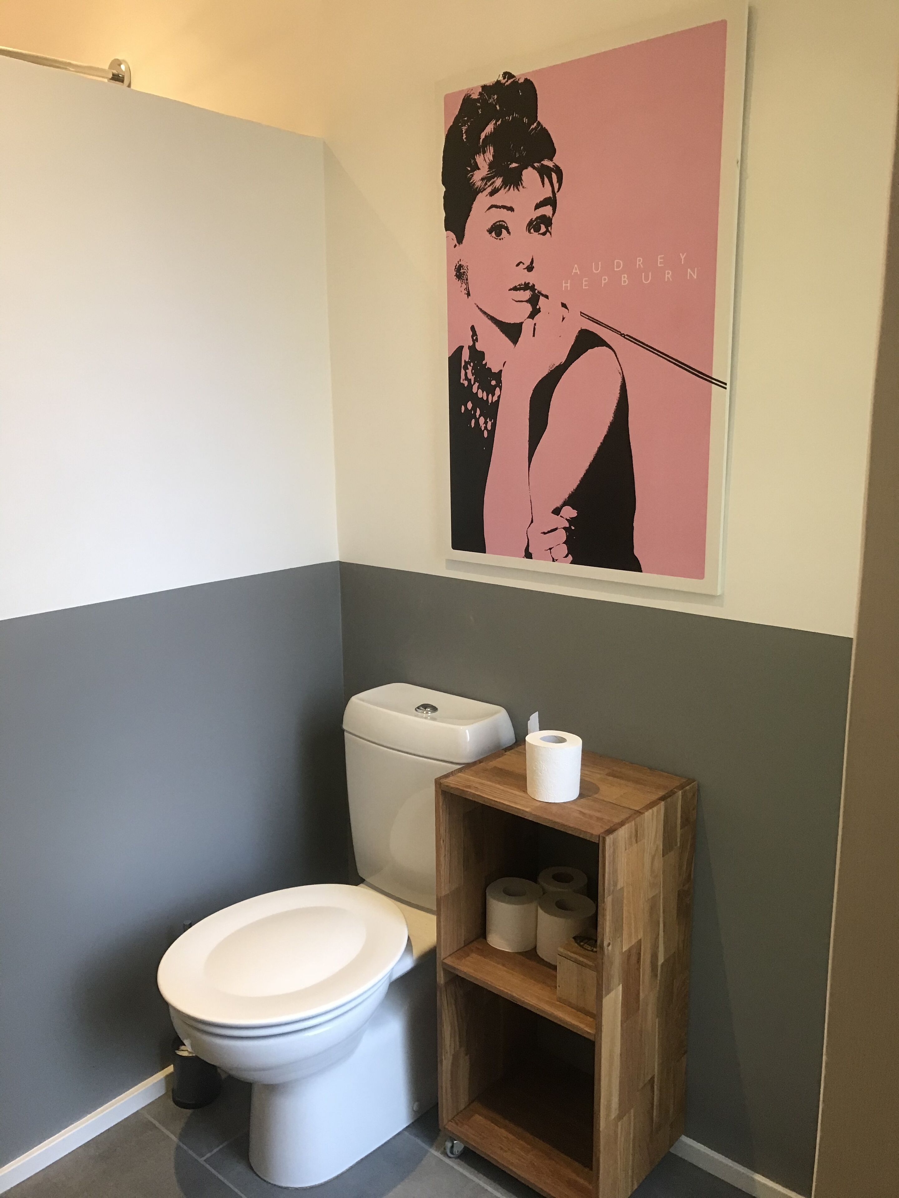 Ein Badezimmer mit Toilette, einem Holzregal mit Toilettenpapier und einem rosa und schwarzen Poster an der Wand.
