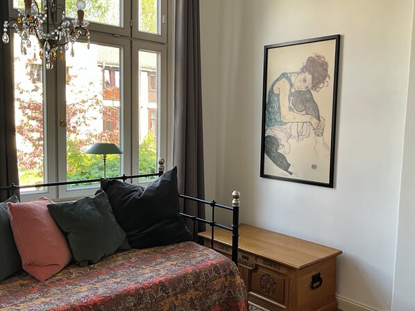 A bedroom with a bed, a nightstand, a wooden chest, a window with curtains, and a chandelier.