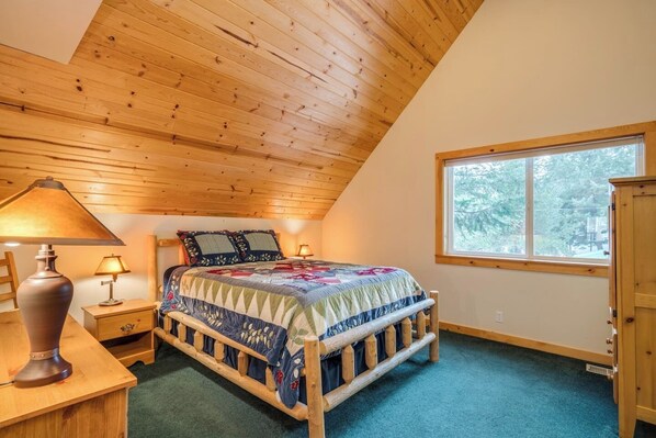 A bedroom with a wooden ceiling, a bed with a quilt, two bedside lamps, a window, and a wooden cabinet. - River views, Sleeps 6, with hot tub & fireplace & game room. (Leavenworth)